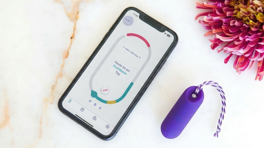 Draufsicht auf ein Smartphone mit der trackle App, die den aktuellen Fruchtbarkeitsstatus anzeigt, daneben liegt der violette trackle Sensor auf einer hellen Marmoroberfläche mit einer rosa Blüte im Hintergrund.