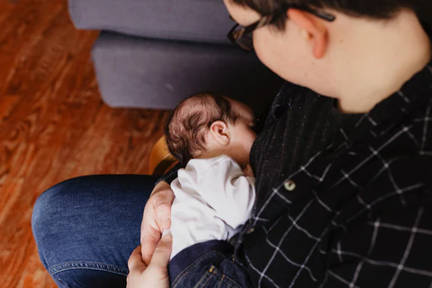 Eine Nahaufnahme aus der Vogelperspektive von einer Mutter, die ihr neugeborenes Baby im Arm hält und stillt. Die Mutter trägt ein dunkel kariertes Hemd, das Baby ein weißes Oberteil. Die Szene vermittelt Ruhe und Geborgenheit. Das Bild illustriert das Thema Stillen und die Anwendung der symptothermalen Methode mit trackle in der postnatalen Phase.