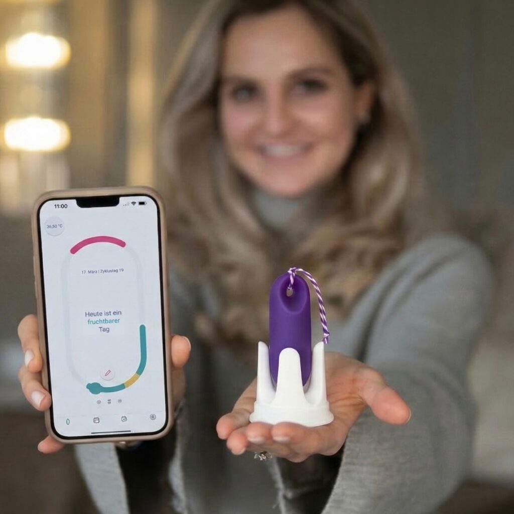 Eine lächelnde Frau hält in der einen Hand ein Smartphone mit der trackle App (Anzeige: ‚Heute ist ein fruchtbarer Tag‘) und in der anderen Hand den violetten trackle Sensor in seiner weißen Halterung.