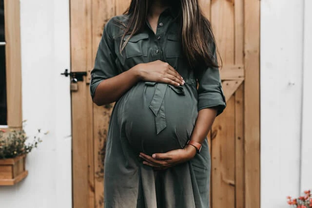 Eine Nahaufnahme einer schwangeren Frau, die ihre Hände liebevoll auf ihren deutlich sichtbaren Babybauch legt. Sie trägt ein olivgrünes Kleid. Im Hintergrund ist eine rustikale Holztür zu sehen. Das Bild symbolisiert Vorfreude, Glück und eine erfolgreiche Familienplanung, die trackle unterstützt.