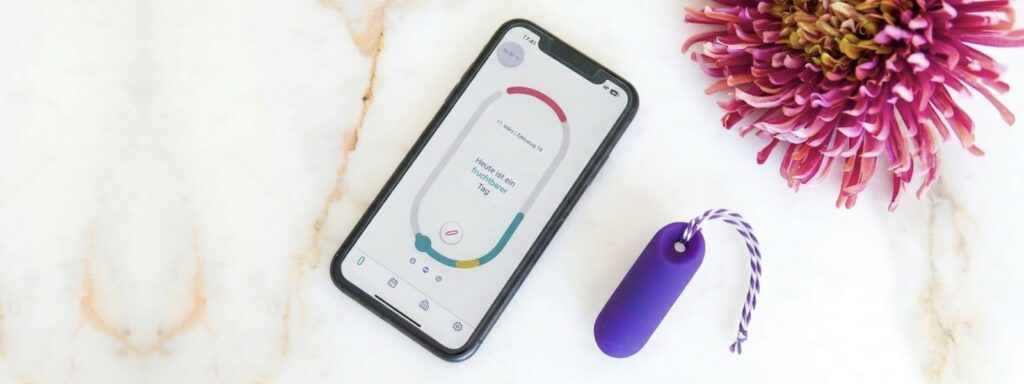 Das trackleCatch Sensorsystem neben einem Smartphone, auf dem die Zyklus-App mit Zyklusdaten und Fruchtbarkeitsanzeige zu sehen ist. Daneben liegt eine pinkfarbene Hortensienblüte auf hellem Marmorhintergrund.