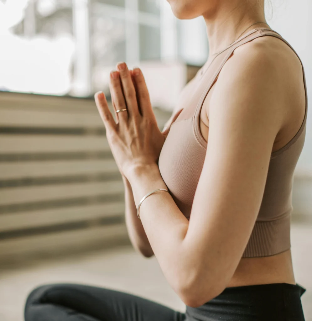 Seitenansicht einer Frau im Yoga-Sitz, die die Hände vor der Brust zum Gruß (Namaste) zusammengelegt hat. Sie trägt ein beigefarbenes Sport-Top.